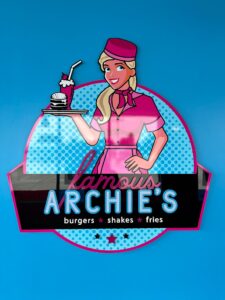 Retro “Famous Archie’s” diner graphic with a waitress in pink, holding a burger and shake, incorporating flat cut acrylic letters as part of the vibrant commercial interior signage.