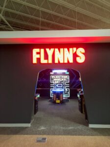 Neon “FLYNN'S” sign above a retro arcade entrance with a vintage “Pac-Man” game, integrating illuminated signage and sign design trends for a nostalgic feel.