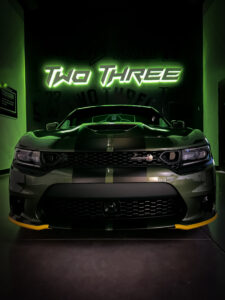 Military-green Dodge Charger in a showroom with “Two Three” wall graphics in a modern style, featuring lit formed plastic dimensional letters. 