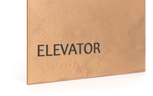 Bronze ADA-compliant elevator sign with tactile Braille and raised text for accessibility.